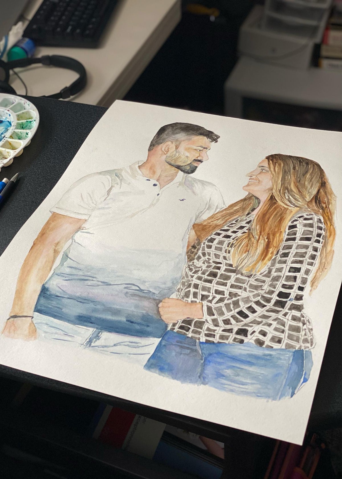 Custom couple portrait being painted in watercolour on a desk, a man and woman embracing beside a paint palette